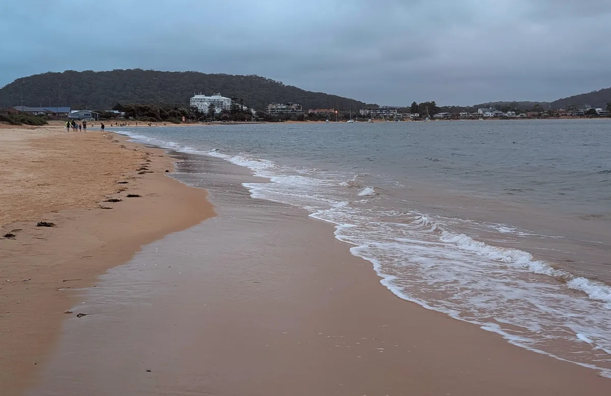Wide view along Ettalong Beach with buildings and a headland beyond