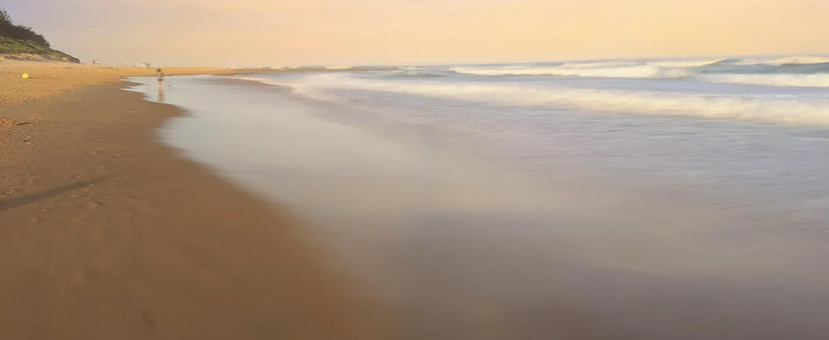 Hazy pastel sunset over the shoreline with a lone walker on the sand